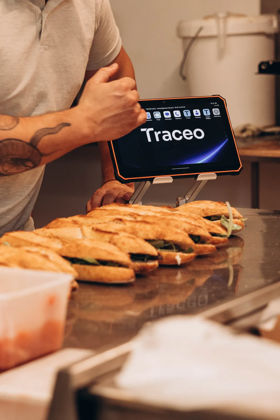 Tablette Traceo sur un support derrière une rangée de sandwichs en préparation dans une boulangerie.
